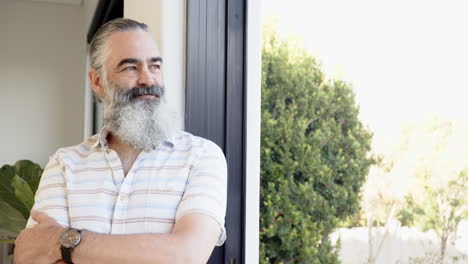 smiling man with beard standing by window, enjoying natural light and greenery, copy space