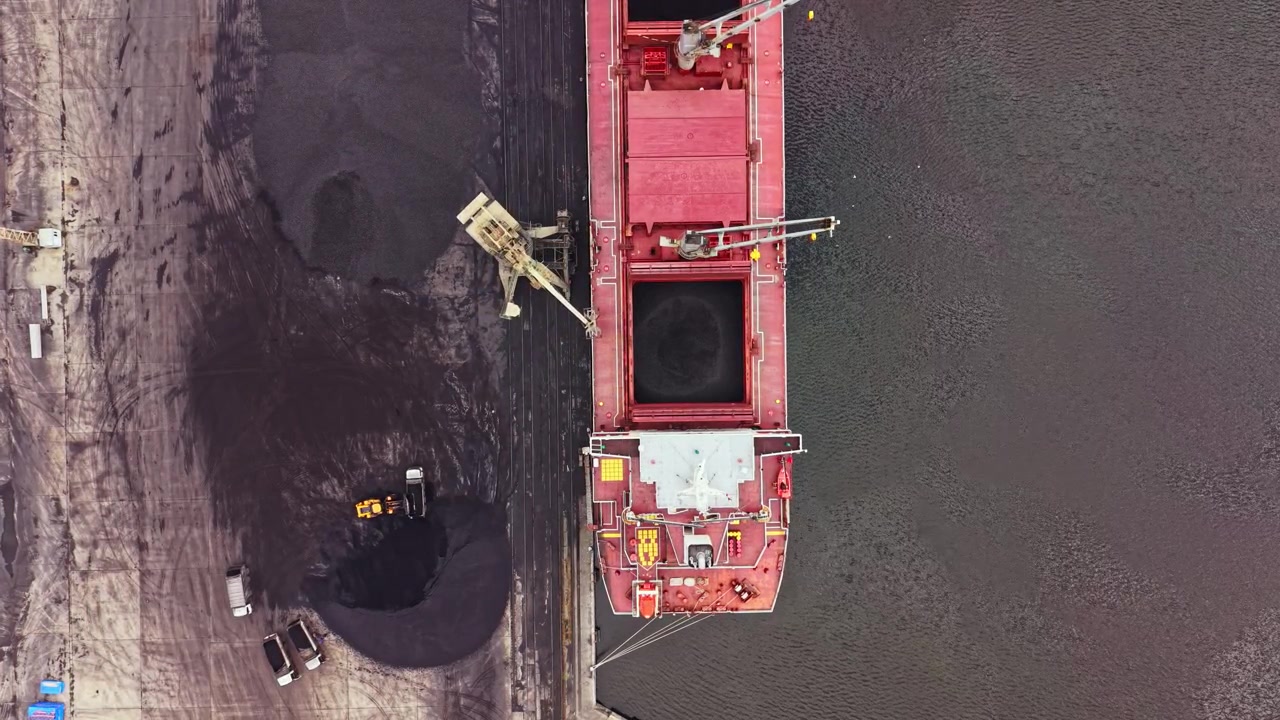 Grande nave da carico che carica carbone in un molo vicino a un fiume durante le ore diurne, con attrezzature che movimentano materiale e veicoli parcheggiati nelle vicinanze.