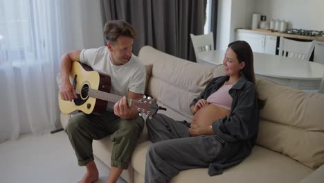 happy middle aged man in white t-shirt playing guitar while hanging out with his pregnant brunette wife at home on sofa during weekend