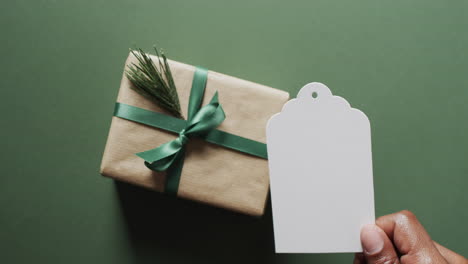 video of christmas presents and hand holding white tag with copy space on green background