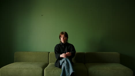 static shot of young woman walking in and sitting down on a green couch in minimalist interior with moody lighting, thinking and pondering in colorful and modern space, beautiful model