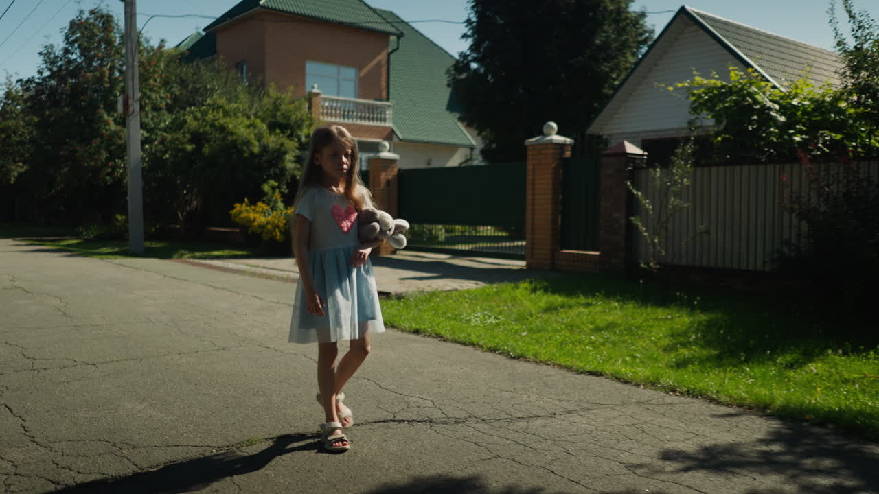 Young girl in light blue gown walks alone on cracked tarred road in residential area, holding stuffed toy tightly while bright sunlight casts shadow on pavement near house and green lawn