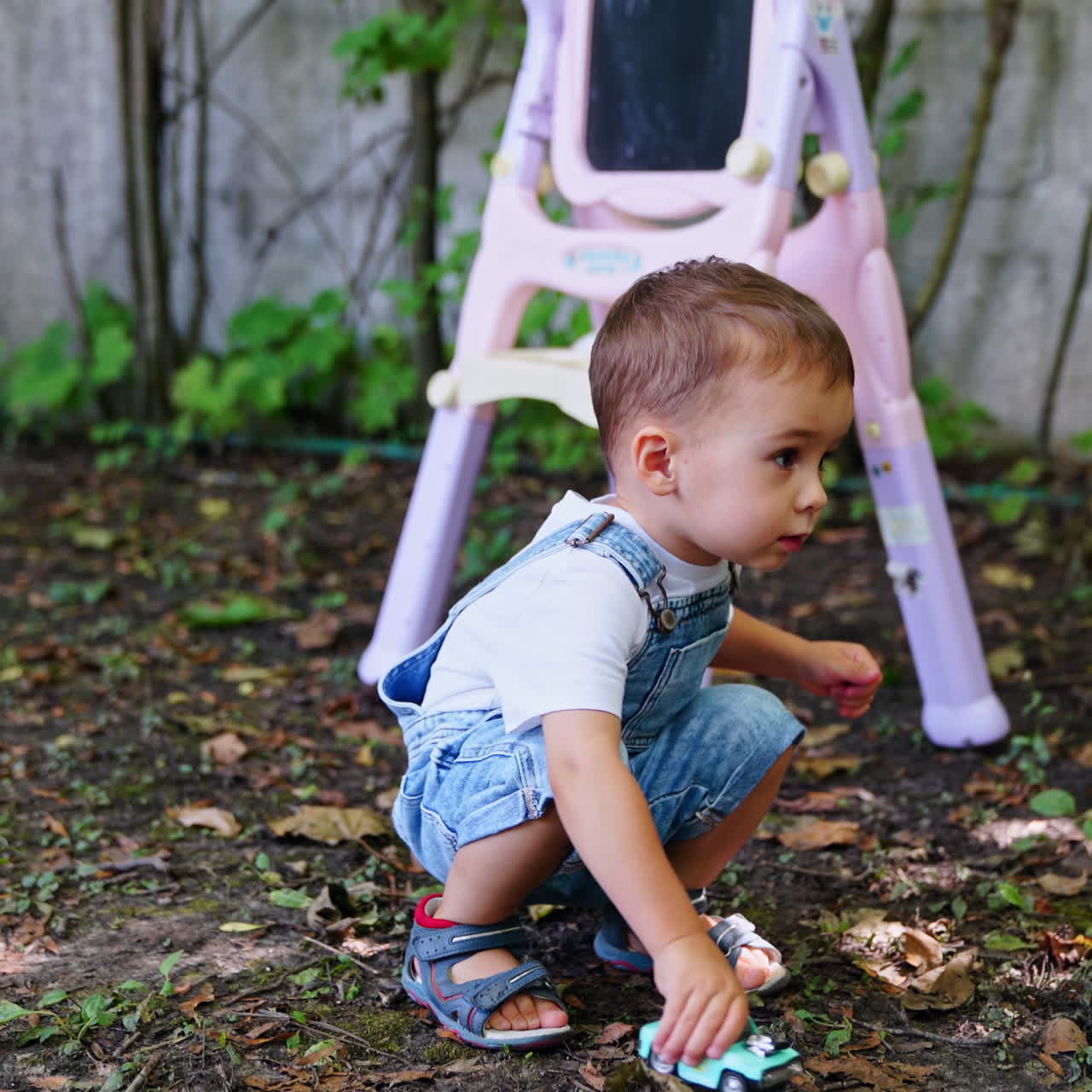 Charmanter kaukasischer Junge im Jeans-Strampler spielt draußen mit einem Spielzeugauto. Kindertafel im Hintergrund.