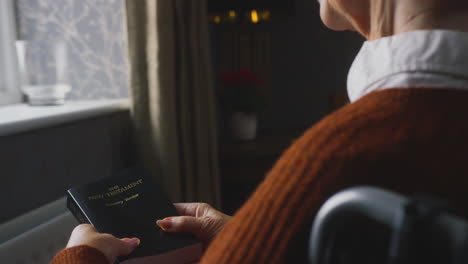 senior woman in wheelchair with bible keeping warm by radiator at home in cost of living crisis