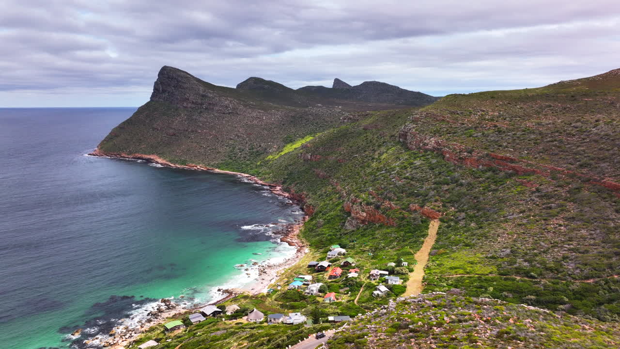 South Africa small beach town Smitswinkel Bay Cape of Good Hope Simon's Town aerial drone cinematic homes during green lush spring summer deep aqua blue ocean sea cloudy mountain cliffs forward motion