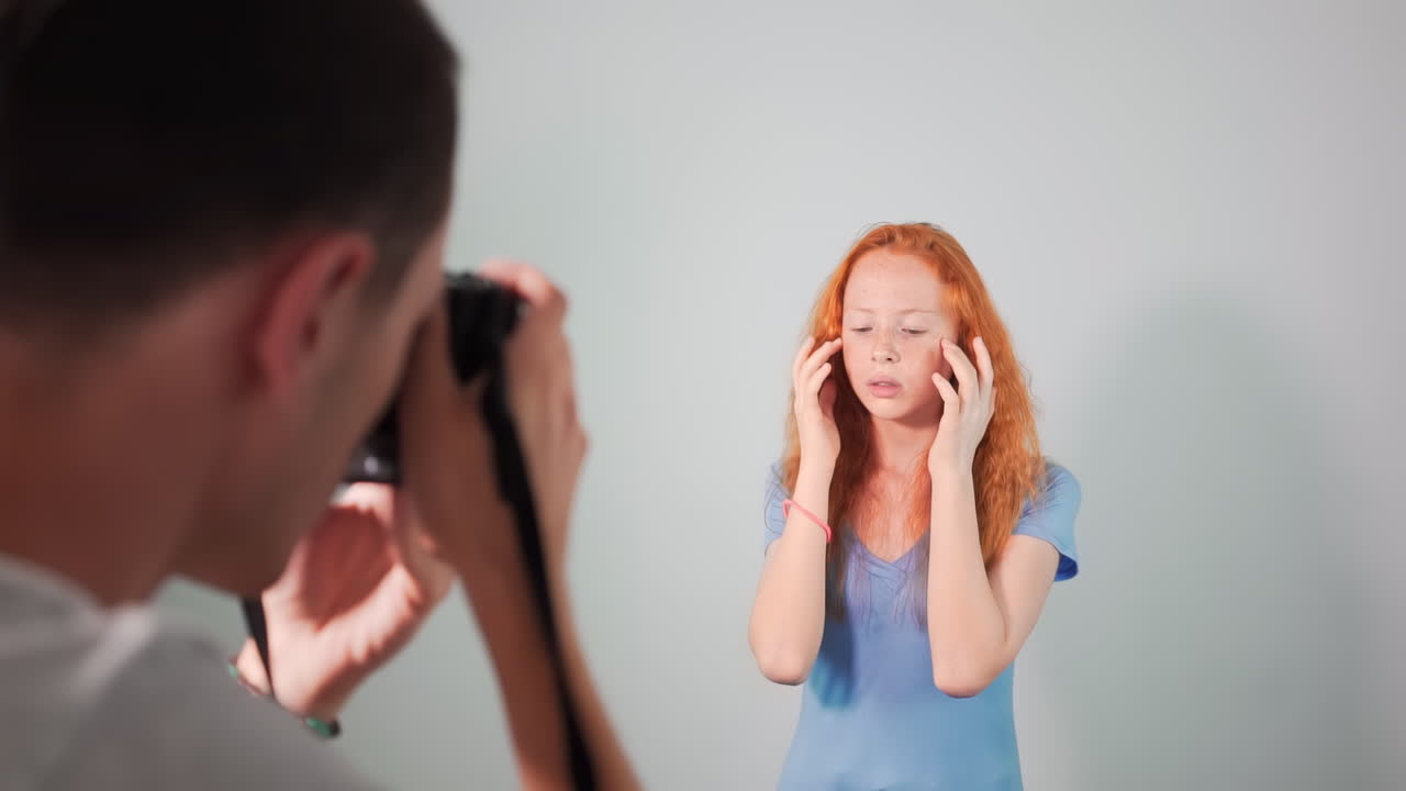Young professional photographer taking photos of a red haired girl in a studio with white background. Rear view. Girl with lots of freckles on the face
