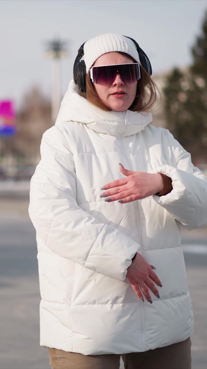 Woman wearing white puffer jacket black headphones and sunglasses moves through outdoor urban setting with snow on ground. Her hands gesture as woman walks or dances in public space