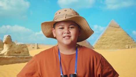 asian boy with a hat and binoculars smiling to camera with arms akimbo while traveling in giza pyramid. boy researcher examines something, travel tourism adventure, close up
