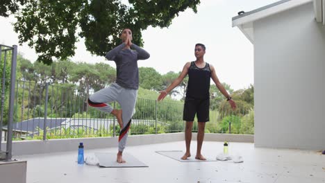 Mixed-race-gay-male-couple-standing-on-terrace-practicing-yoga