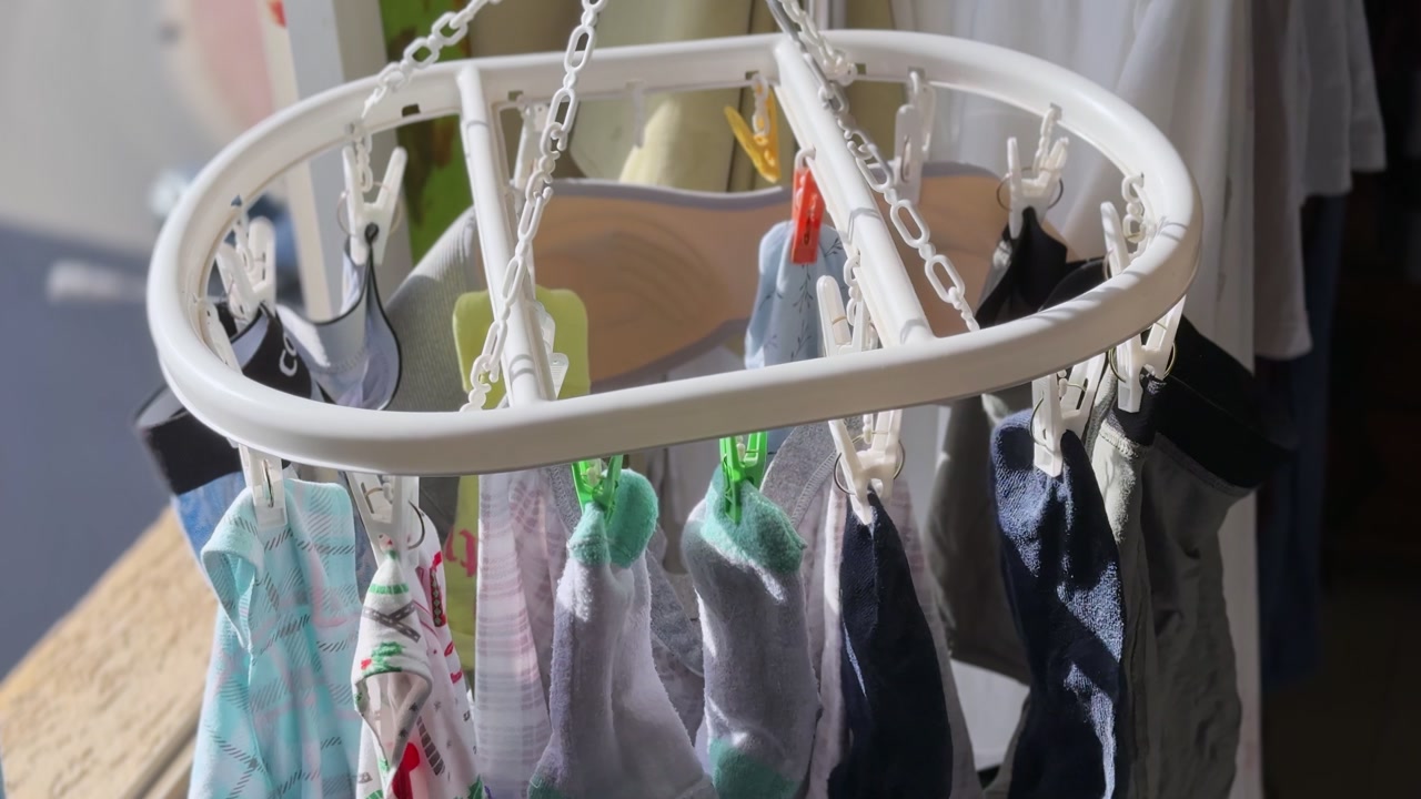 Close-up of assorted socks and small garments clipped to a white drying rack beside a sunny window, showing everyday household laundry care inside a bright apartment.