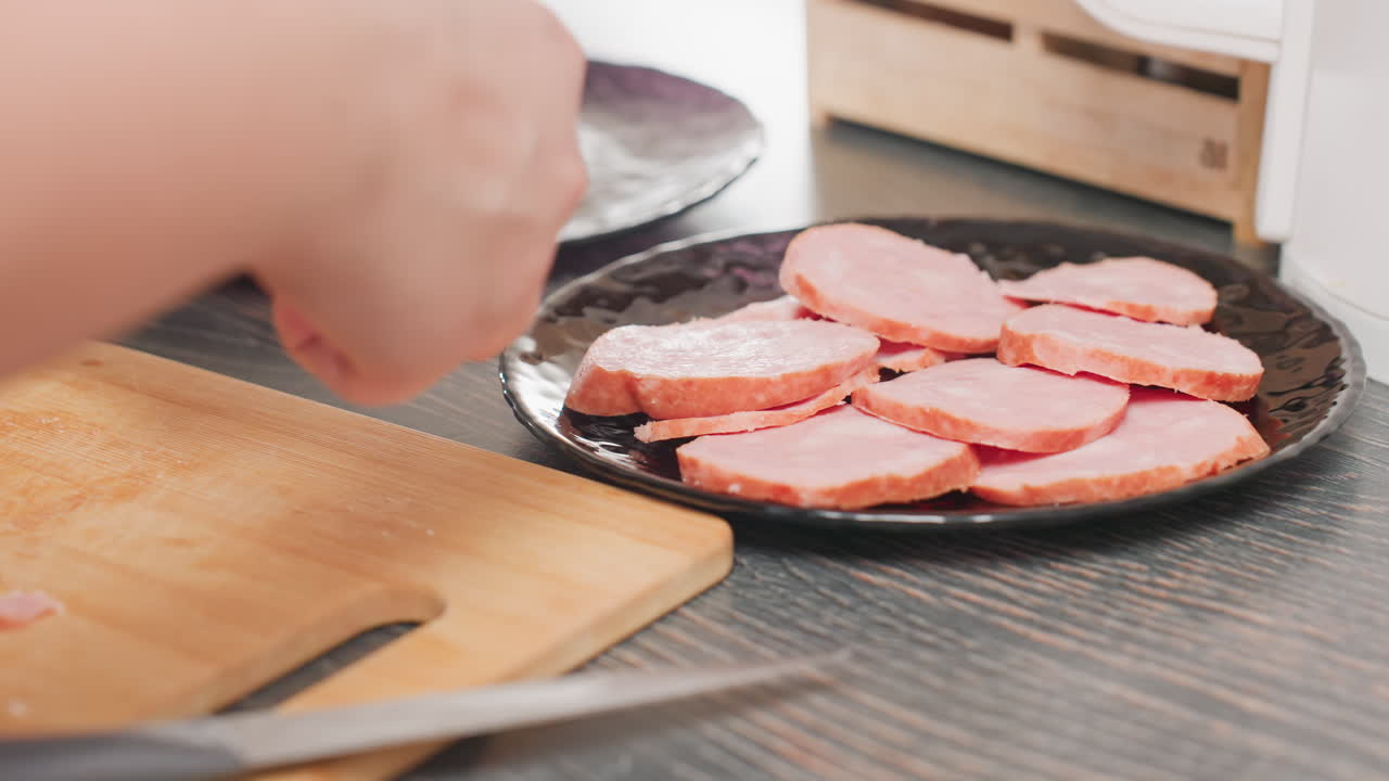 close up hand view of person arranging round sausage slices on black plate beside wooden cutting board, freshly cut meat slices visible, knife resting nearby, kitchen food preparation scene