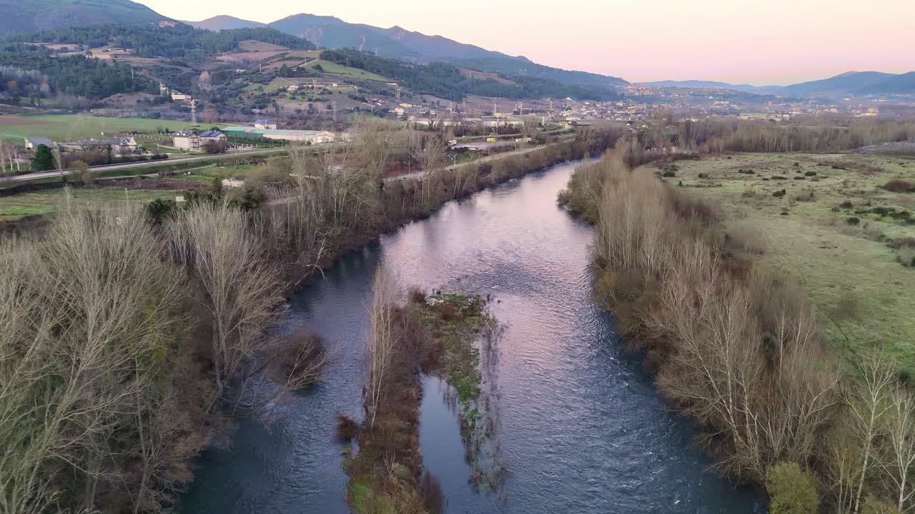Aerial shot of the Sil River flowing through a rural landscape with trees, fields, and distant mountains under a colorful sunset sky.
