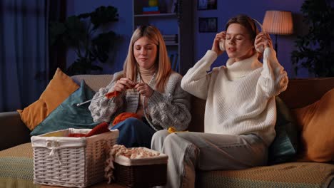 Creative Knitting Evening With Girlfriends on a Cozy Night. Two women enjoying a relaxing evening at home, knitting and laughing on the couch. The friends watch a video lesson on the laptop and learn to knit new patterns with needles