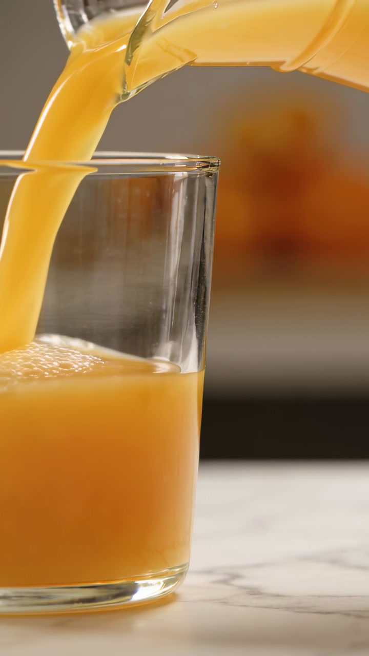 Vertical video: Tilting pitcher pouring orange juice into clear glass on marble top for serving