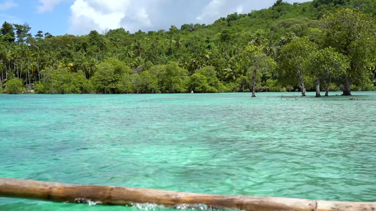 POV looking out from a small fishing boat sailing around the Banggai Islands in Indonesia. You can see the crystal clear blue ocean water and the dense green jungle on the nearby islands