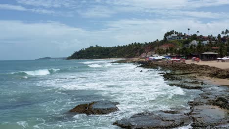 Dolly-in-aerial-drone-shot-of-the-tropical-Tibau-do-Sul-peninsula-with-waves-crashing-into-algae-covered-rocks,-tourists-enjoying-shade-under-colorful-umbrellas-in-Rio-Grande-do-Norte,-Brazil