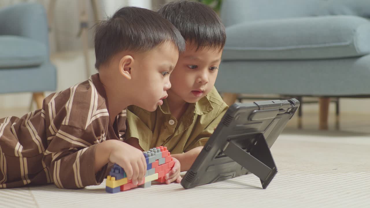 플라스틱 장난감 벽돌을 가진 아시아 어린이들이 바닥에 누워서 집에서 함께 태블릿에서 영화를 보고 있습니다.