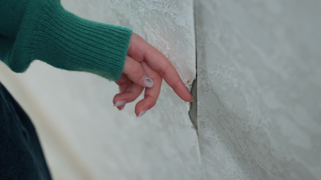 Nærbillede af afskallende puds og hårfin revne i smal gang, kvinde i grøn sweater rækker ud med fingerspidsen for at vurdere skaden, dæmpet lys, tekstureret overflade, eftertænksom stemning, planlægger reparation