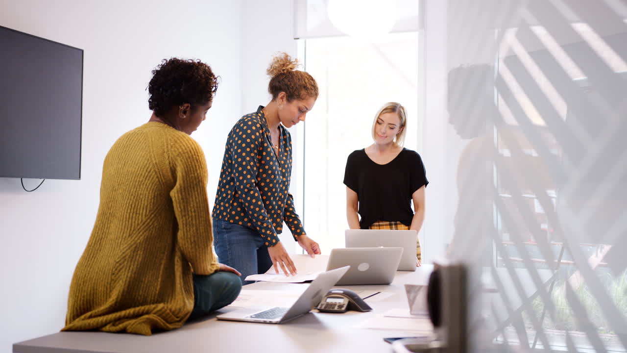 Three serious female creatives discussing paperwork during a casual meeting in an office meeting room, seen from doorway