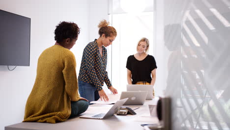 three serious female creatives discussing paperwork during a casual meeting in an office meeting room, seen from doorway