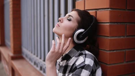 The-woman-pensively-listens-to-the-music-in-the-headphones,-leaning-on-the-wall-of-the-fence-in-the-street.-My-eyes-are-closed.-Make-up.-Close-up