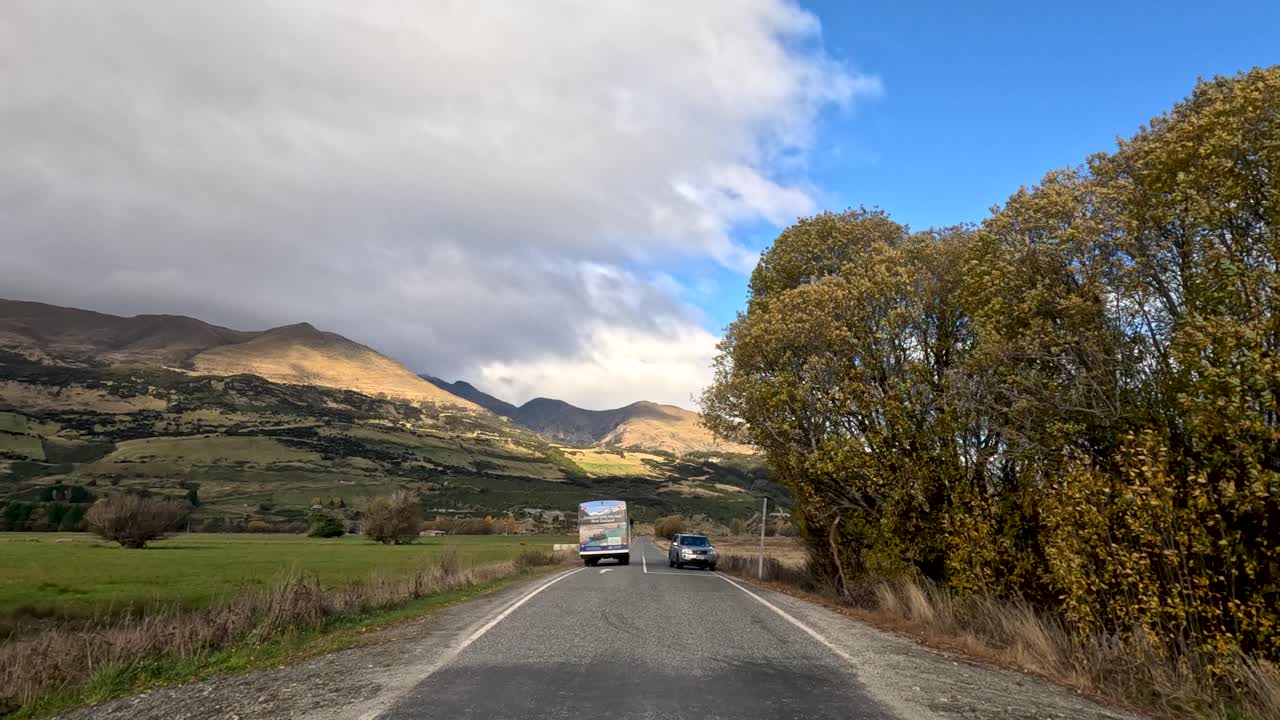 Vehicle follows truck over rural bridge through scenic valley under partly cloudy sky, steady motion