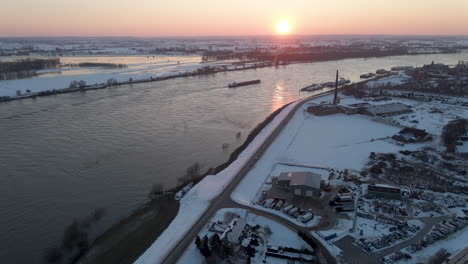 Aerial-jib-up-of-cargo-ship-on-river-with-a-setting-sun-in-the-background-in-winter---wide