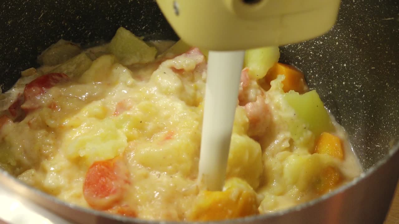 Close-up of a creamy vegetable soup being blended in a pot, showing chunks of carrots, potatoes, and other vegetables turning into a smooth, rich mixture