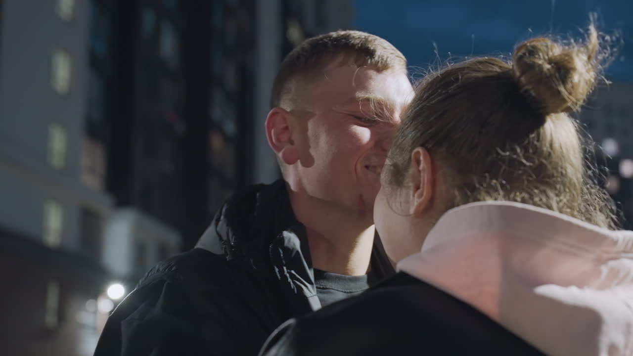 Close up of man laughing warmly while giving his lady gentle peck on cheek under dusk sky with blurred lights from tall city buildings creating tender and joyful moment in urban evening setting