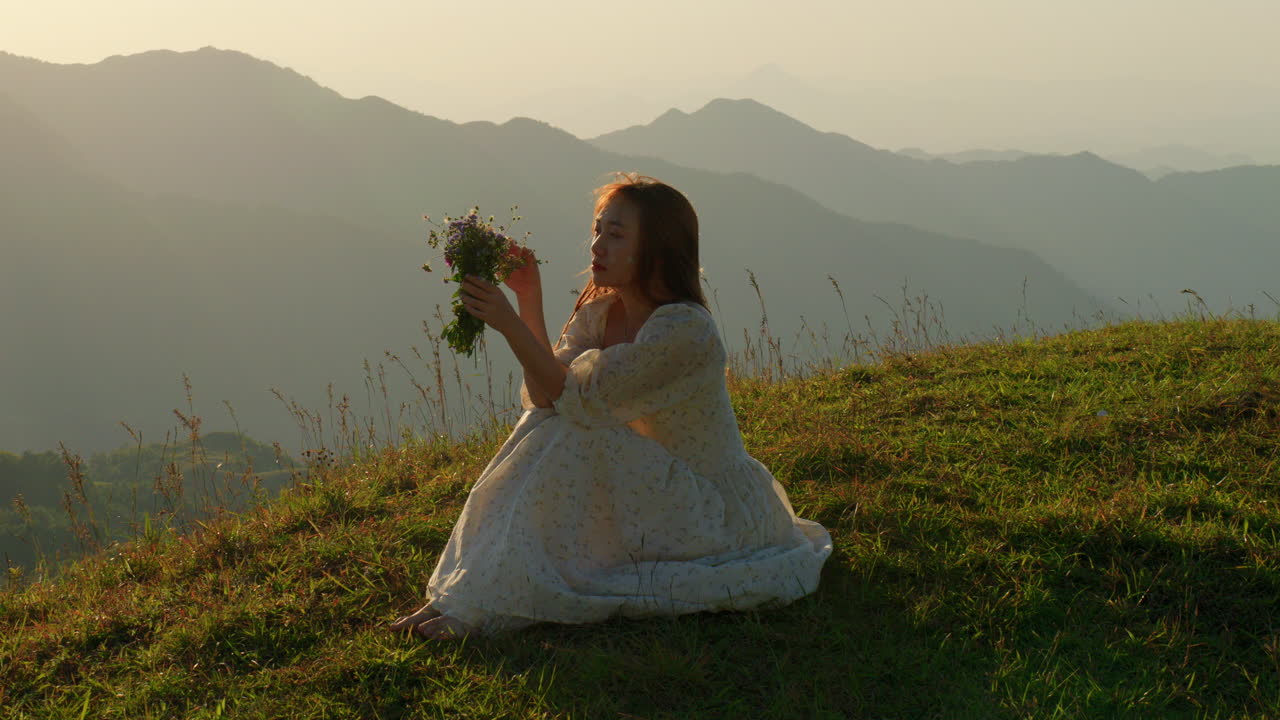 lonely asiatic woman sitting in nature at sunset with mountain landscape holding a bouquet of flowers