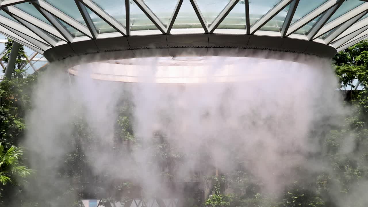 Dense mist rapidly ascends from a large indoor waterfall beneath a geometric glass roof, surrounded by lush greenery and natural daylight, captured in a stationary wide shot
