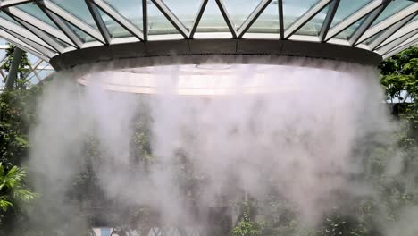 dense mist rapidly ascends from a large indoor waterfall beneath a geometric glass roof, surrounded by lush greenery and natural daylight, captured in a stationary wide shot