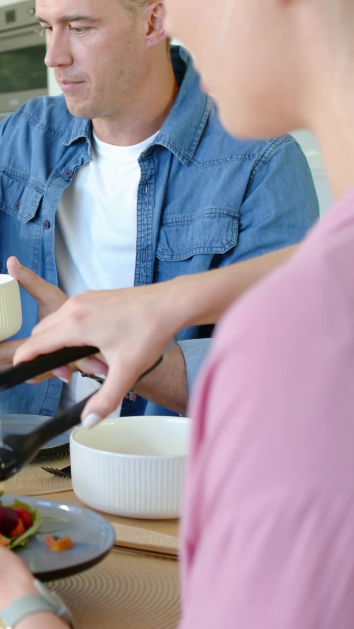 Vertical video: Serving woman in pink top with black tongs as meal start, man holding bowl at home