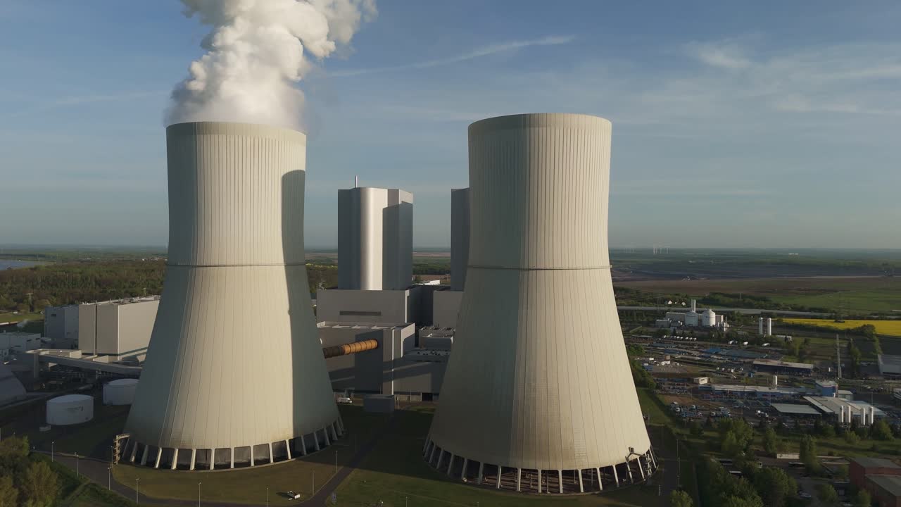 Drone flies towards the front of the Lippendorf power plant cooling towers near Leipzig, Germany. Massive infrastructure with steam and clear blue sky on a spring day.