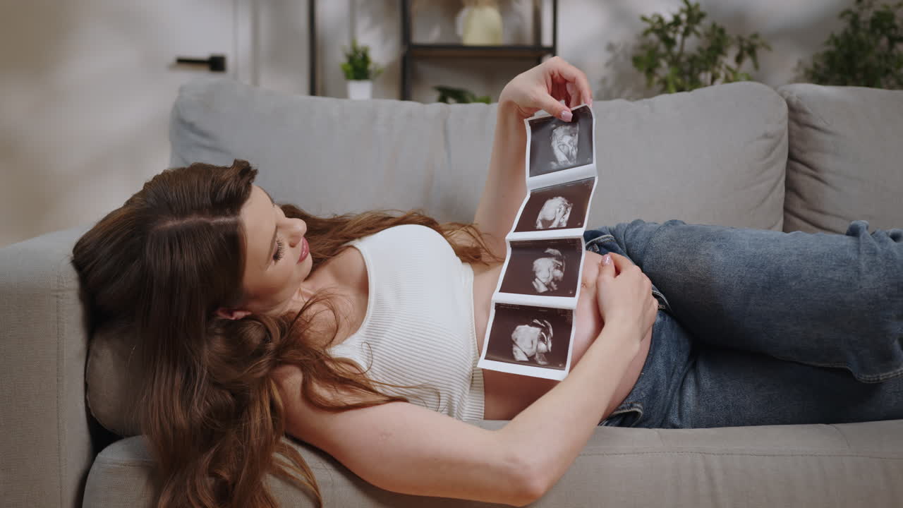 Expecting mother reclines on sofa holding ultrasound images showing developing fetus anticipating