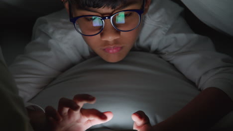 close up de um menino no quarto em casa usando telefone celular para mensagem de texto sob coberturas ou edredom à noite 1