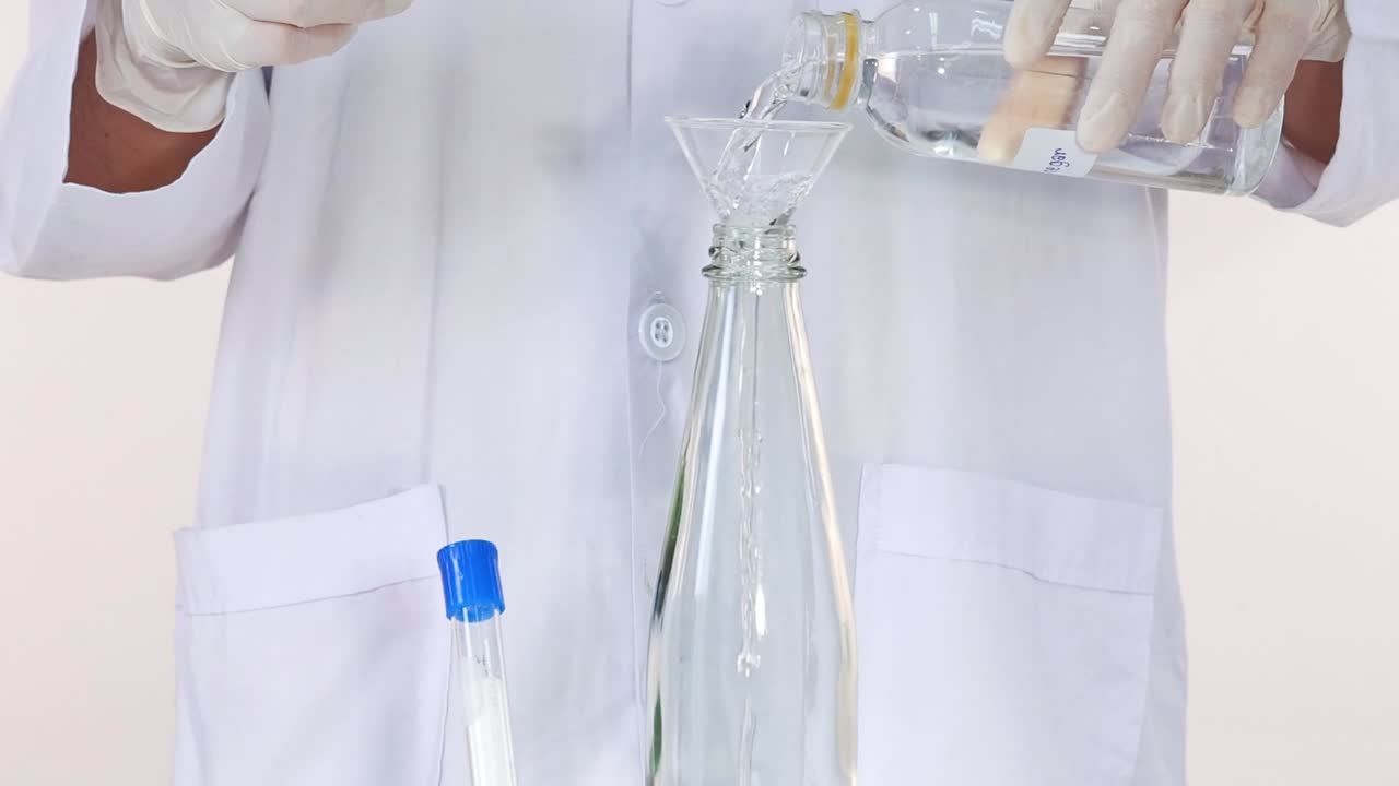 A scientist in a lab coat pours liquid into a funnel over a glass bottle.