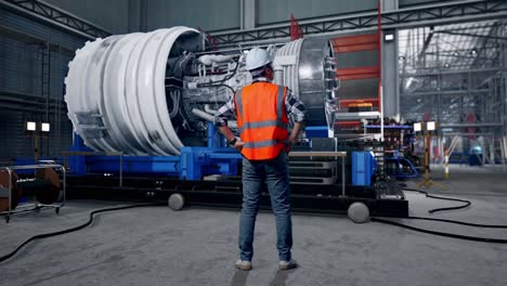 full body back view of a male engineer wearing safety helmet looking around while standing with arms akimbo with airplane engine maintenance conducted, analytics checking the turbine