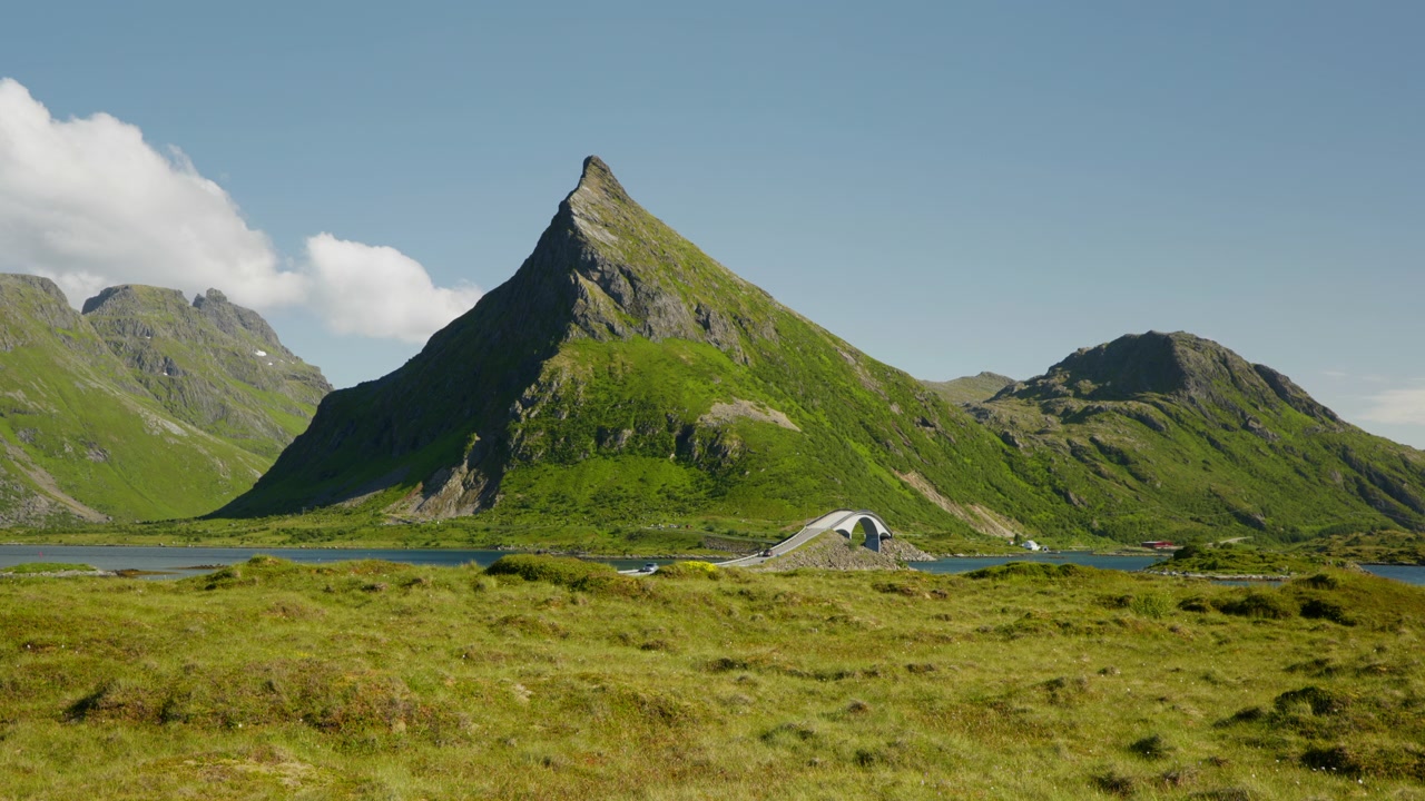 En takket bjergtop, en buet bro og en blå fjord ligger bag åben græsklædt tundra under klar sommerhimmel