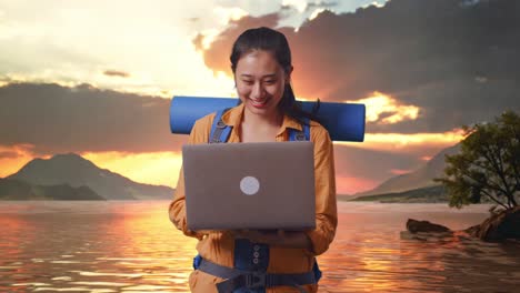 excursionista femenina asiática con mochila de montañismo usando una computadora portátil mientras está de pie en un lago durante la hora del atardecer