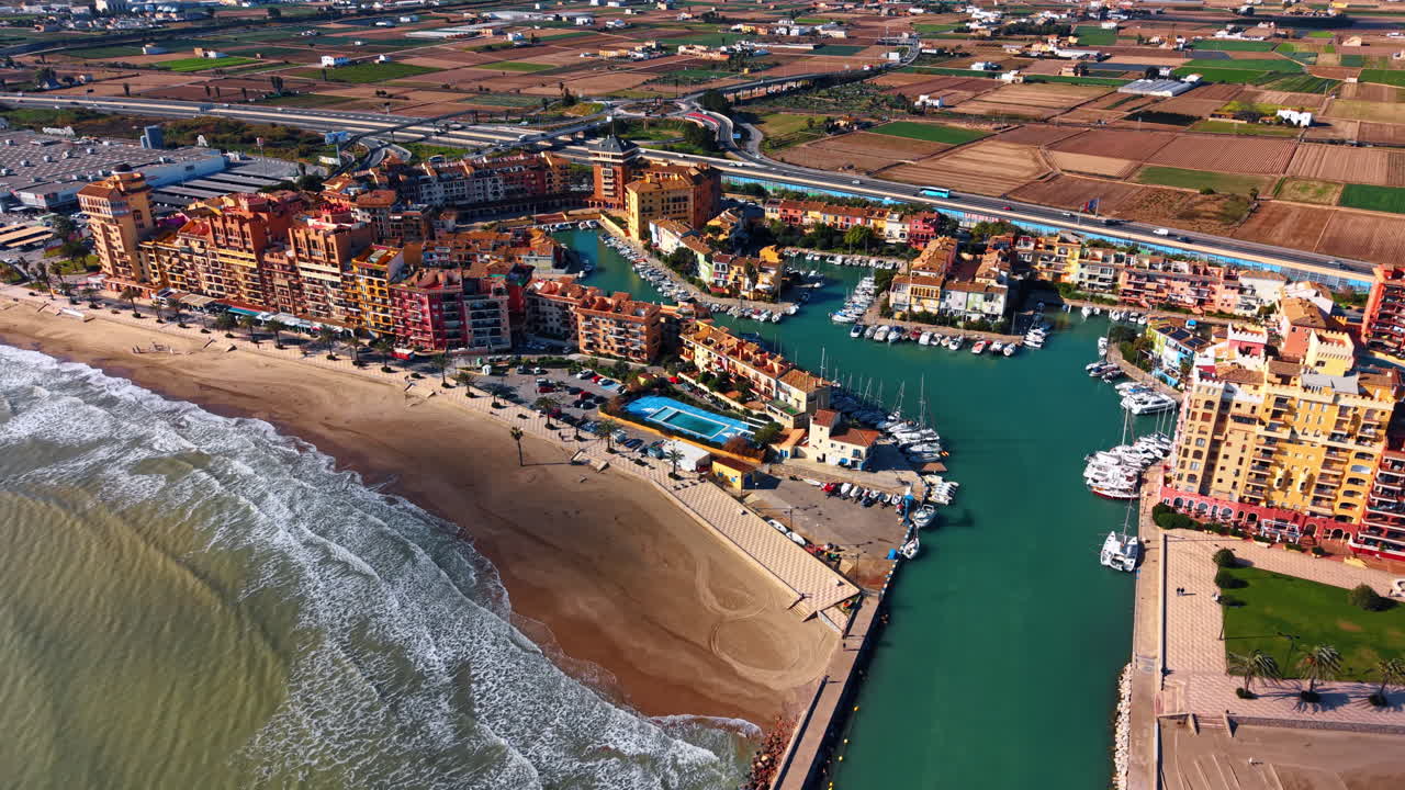 Marina mediterranea e spiaggia costiera. Ampia vista su condomini colorati, yacht, litorale sabbioso e montagne lontane sotto un cielo sereno.