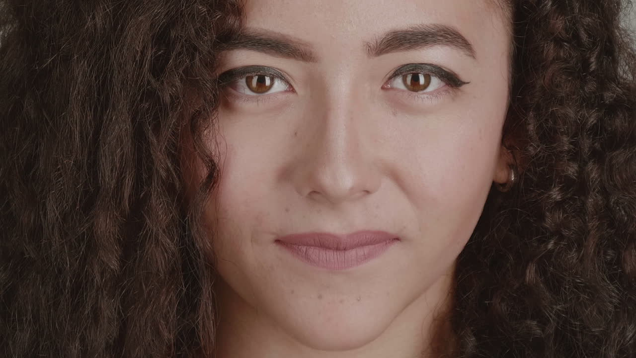 High-quality 4K footage of a confident young multi-ethnic woman with beautiful curly hair. She looks directly into the lens with a subtle, smug smile against a simple grey background