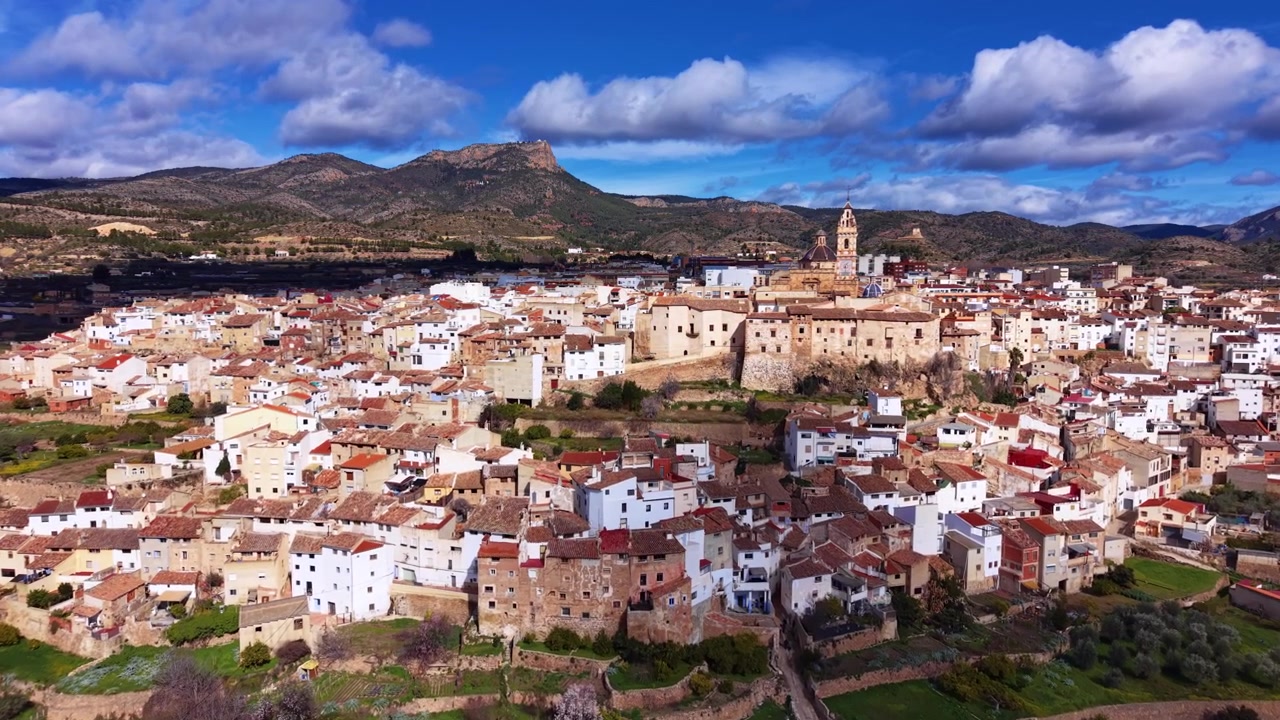 Panoramaudsigt over Chelva, en traditionel spansk landsby i Valencia-regionen, med hvide og terrakottafarvede tage, et historisk kirketårn og bjergkulisse under blå himmel
