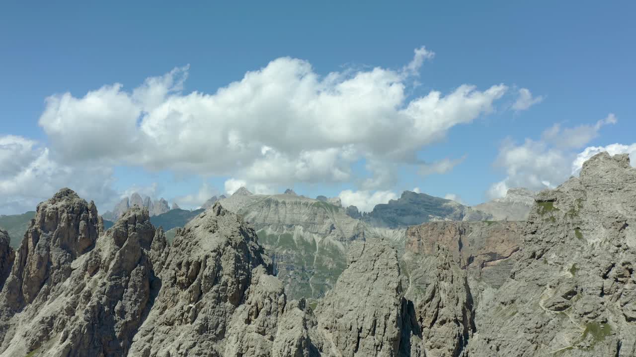 Aerial drone view capturing jagged limestone peaks and rugged terrain under a bright blue sky with scattered clouds in the Dolomites region of Italy