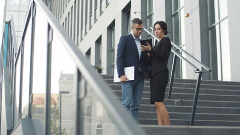 Vista-Lejana-De-Una-Mujer-De-Negocios-Mostrando-Algo-A-Un-Hombre-De-Negocios-En-Un-Dispositivo-De-Tableta-Mientras-Están-Parados-En-Los-Escalones