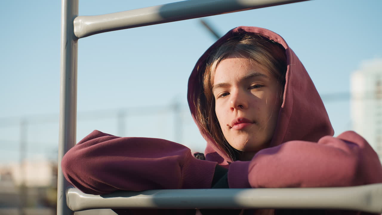 Portrait of calm focused young woman resting on exercise bar during outdoor break under sunlight, wearing hoodie and vest. Urban fitness scene captures moment of relaxation and concentration