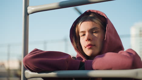 portrait of calm focused young woman resting on exercise bar during outdoor break under sunlight, wearing hoodie and vest. urban fitness scene captures moment of relaxation and concentration