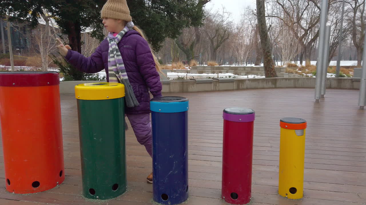 Una ripresa cinematografica al rallentatore cattura una bambina con una giacca invernale viola e un cappello che suona colorati tamburi a percussione nel giardino musicale all'aperto della Casa della Musica Ungherese.