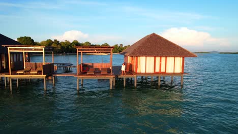 Mujer-Caminando-En-Un-Muelle-De-Madera-Cerca-De-Una-Villa-Sobre-El-Agua-En-Medio-Del-Mar-Al-Amanecer,-Drones-Volando-Hacia-Atrás-En-La-Isla-Leebong-En-Belitung-Indonesia