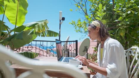 side view young female businesswoman using laptop outdoors at terrace with palm trees, remote online working during summer vacation